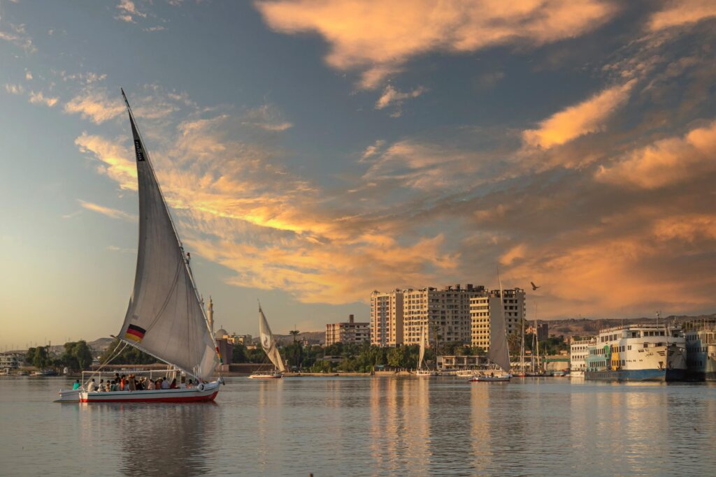 A traditional felucca sails the Nile at sunset with cityscape in the background.